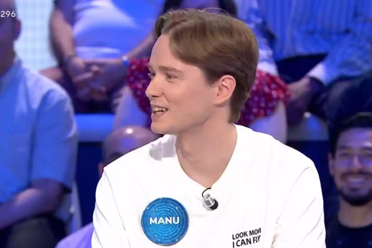 Un jove amb samarreta blanca i una placa blava que diu Manu participa en un programa de televisió amb públic al fons. Un jove amb samarreta blanca i una placa blava que diu Manu participa en un programa de televisió amb públic al fons.