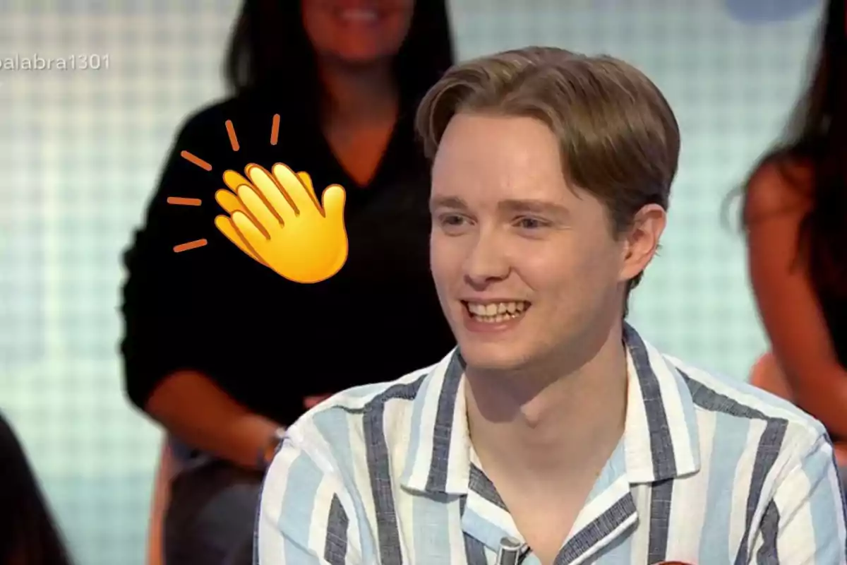 Jove somrient amb camisa de ratlles blaves i blanques en un programa de televisió amb un emoji d’aplaudiments al fons