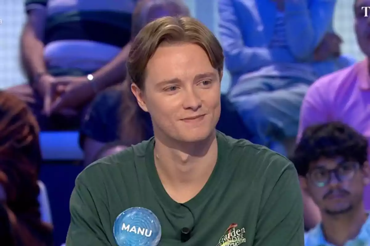 Un jove amb samarreta verda i una placa que diu Manu participa en un programa de televisió amb públic al fons. Un jove amb samarreta verda i una placa que diu Manu participa en un programa de televisió amb públic al fons.