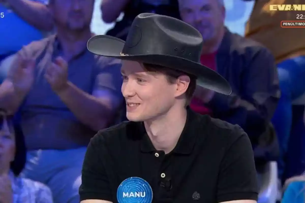 Un jove amb barret negre somriu mentre participa en un programa de televisió, porta una samarreta negra i una placa blava amb el nom Manu, el públic aplaudeix al fons. Un jove amb barret negre somriu mentre participa en un programa de televisió, porta una samarreta negra i una placa blava amb el nom Manu, el públic aplaudeix al fons.
