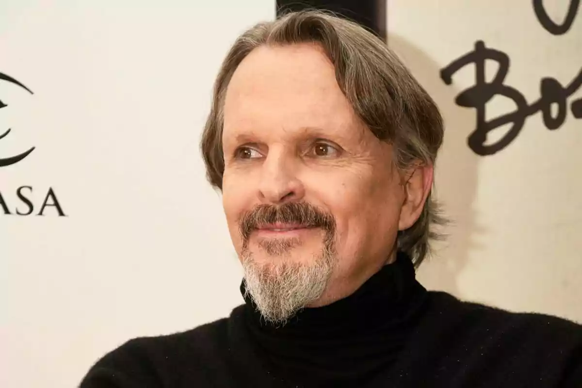 Hombre de mediana edad con barba y bigote canosos, cabello castaño, usando suéter negro y sonriendo levemente frente a un fondo claro con letras negras. Hombre de mediana edad con barba y bigote canosos, cabello castaño, usando suéter negro y sonriendo levemente frente a un fondo claro con letras negras.