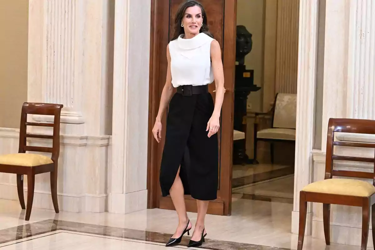 Dona de cabell llarg i solt vestida amb una brusa blanca de tirants i una faldilla negra midi amb cinturó ample, caminant en un interior elegant amb cadires de fusta i parets de marbre. Dona de cabell llarg i solt vestida amb una brusa blanca de tirants i una faldilla negra midi amb cinturó ample, caminant en un interior elegant amb cadires de fusta i parets de marbre.