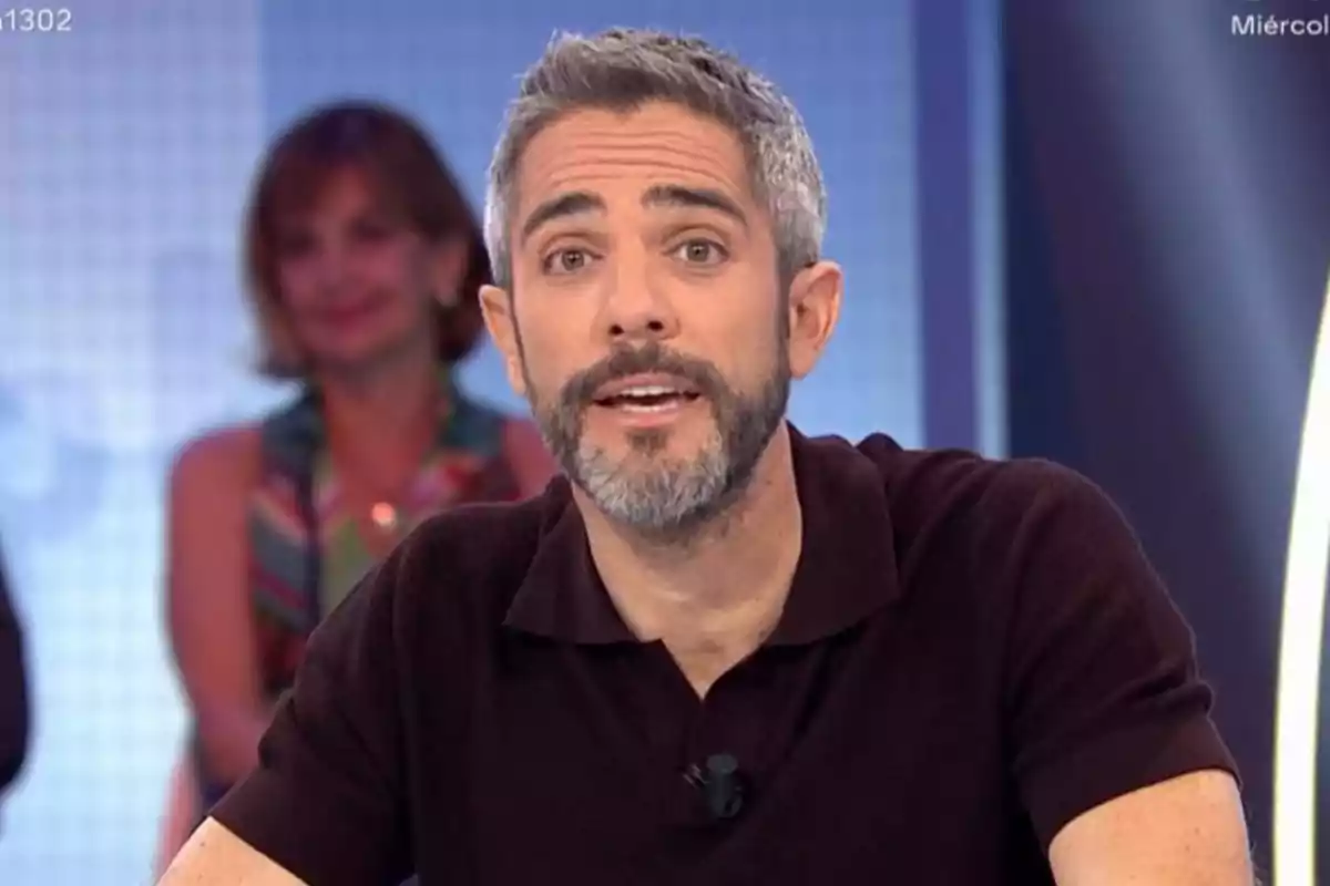 Short-haired, gray-bearded man speaking on a television show with a blurred woman in the background Short-haired, gray-bearded man speaking on a television show with a blurred woman in the background
