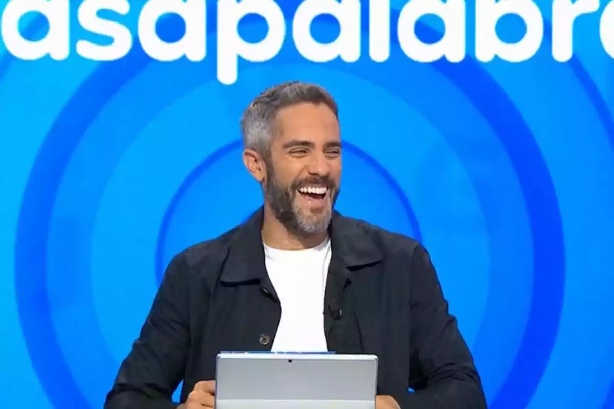 Home de cabell curt i barba somrient mentre sosté una tauleta davant d’un fons blau amb lletres blanques Home de cabell curt i barba somrient mentre sosté una tauleta davant d’un fons blau amb lletres blanques