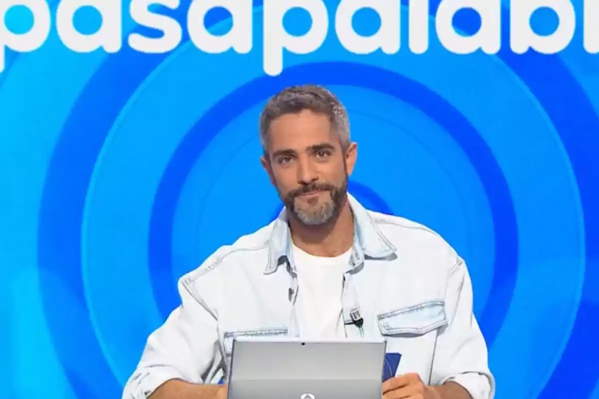 Home de cabells curts i barba assegut davant d’un ordinador portàtil amb un fons blau i lletres blanques d’un programa de televisió Home de cabells curts i barba assegut davant d’un ordinador portàtil amb un fons blau i lletres blanques d’un programa de televisió