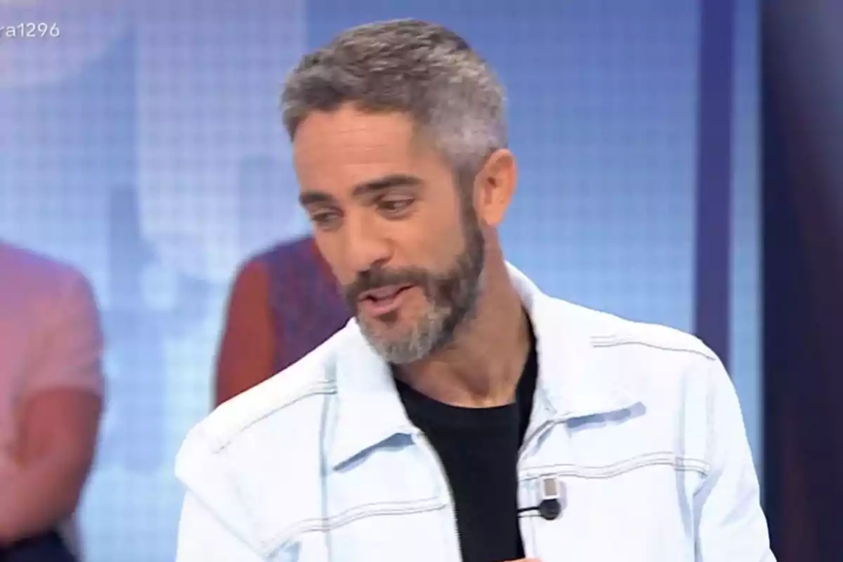 Home de cabell curt i barba amb jaqueta clara i samarreta fosca en un plató de televisió Home de cabell curt i barba amb jaqueta clara i samarreta fosca en un plató de televisió