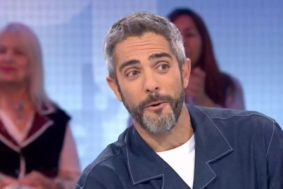 Home de cabells i barba canosos amb expressió de sorpresa en un plató de televisió amb persones desenfocades al fons Home de cabells i barba canosos amb expressió de sorpresa en un plató de televisió amb persones desenfocades al fons