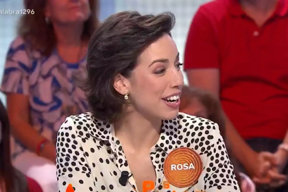 Dona de cabell curt i fosc amb una brusa de punts blancs i negres participant en un programa de televisió amb una placa que diu Rosa Dona de cabell curt i fosc amb una brusa de punts blancs i negres participant en un programa de televisió amb una placa que diu Rosa
