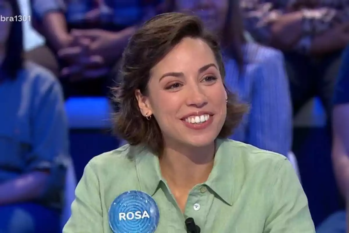 Dona somrient amb cabell curt i camisa verd clar participant en un programa de televisió amb una placa que diu Rosa Dona somrient amb cabell curt i camisa verd clar participant en un programa de televisió amb una placa que diu Rosa