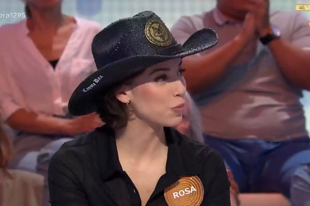 Dona amb barret negre i camisa fosca amb una placa que diu Rosa participant en un programa de televisió amb públic de fons Dona amb barret negre i camisa fosca amb una placa que diu Rosa participant en un programa de televisió amb públic de fons
