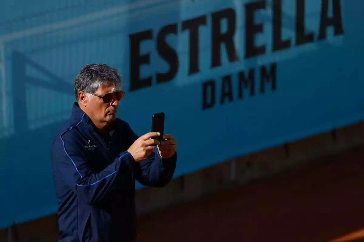 Gray-haired man wearing sunglasses taking a photo with his cellphone in front of a blue sign that says Estrella Damm Gray-haired man wearing sunglasses taking a photo with his cellphone in front of a blue sign that says Estrella Damm
