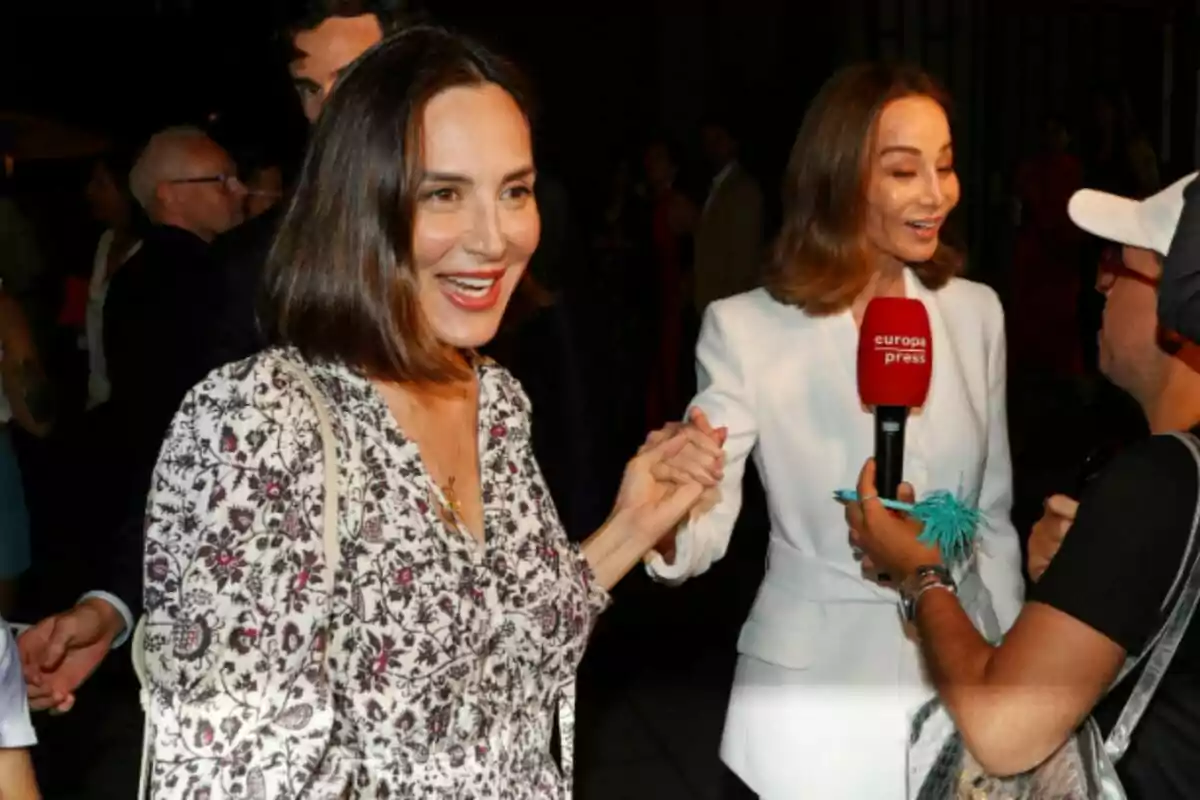 Isabel Preysler i Tamara Falcó, somrients, es saluden i es donen la mà en un esdeveniment mentre una d’elles és entrevistada per un reporter que sosté un micròfon vermell d’Europa Press, envoltades de més persones en un ambient nocturn.