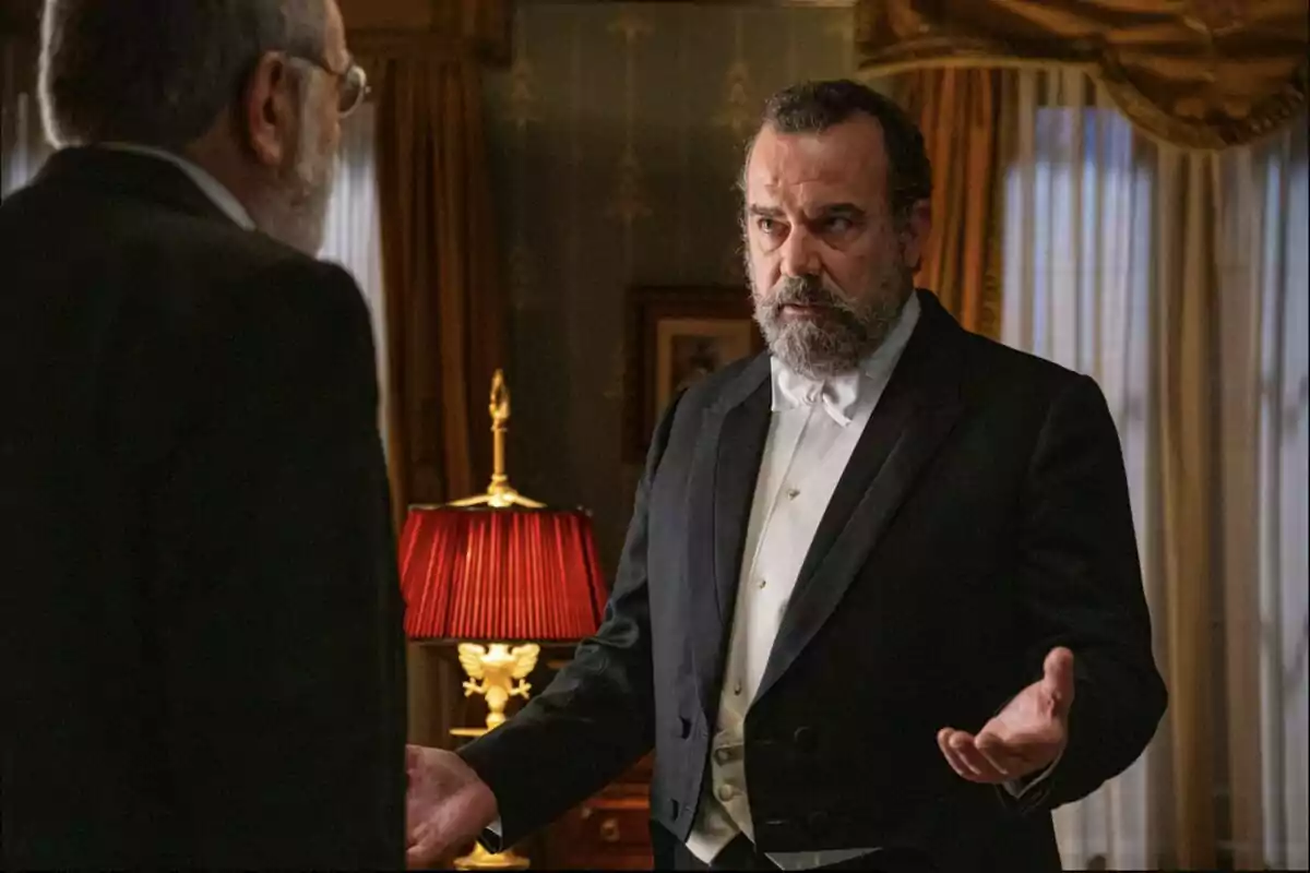 Alonso amb barba i esmòquing blanc i negre gesticulant amb les mans mentre parla amb Ròmul en una habitació elegant amb cortines daurades i una làmpada de pantalla vermella. Captura de 'La Promesa' Alonso amb barba i esmòquing blanc i negre gesticulant amb les mans mentre parla amb Ròmul en una habitació elegant amb cortines daurades i una làmpada de pantalla vermella. Captura de 'La Promesa'
