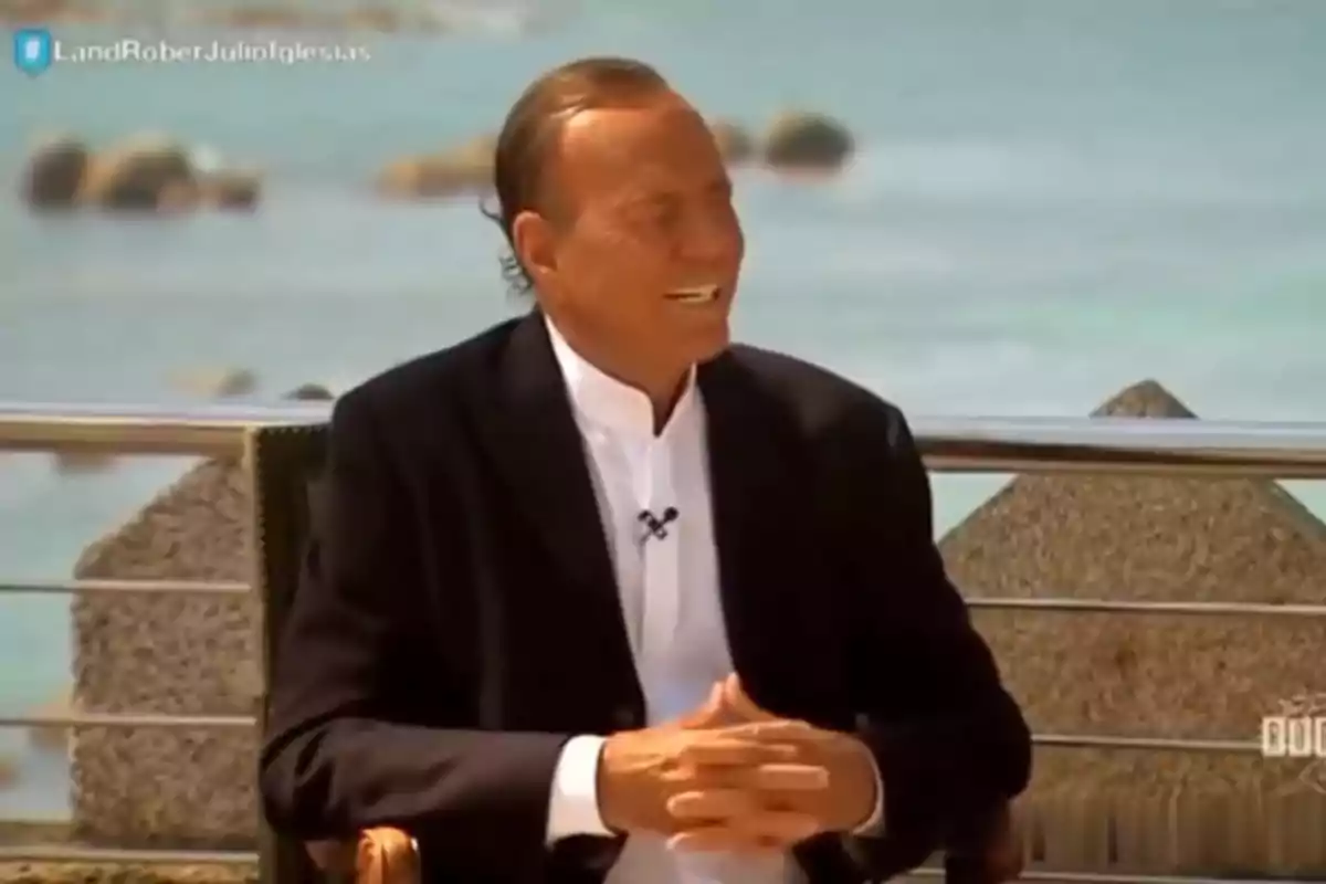 Julio Iglesias in a dark suit and white jersey sitting outdoors by the sea, smiling during an interview. Still from 'Land Rober'