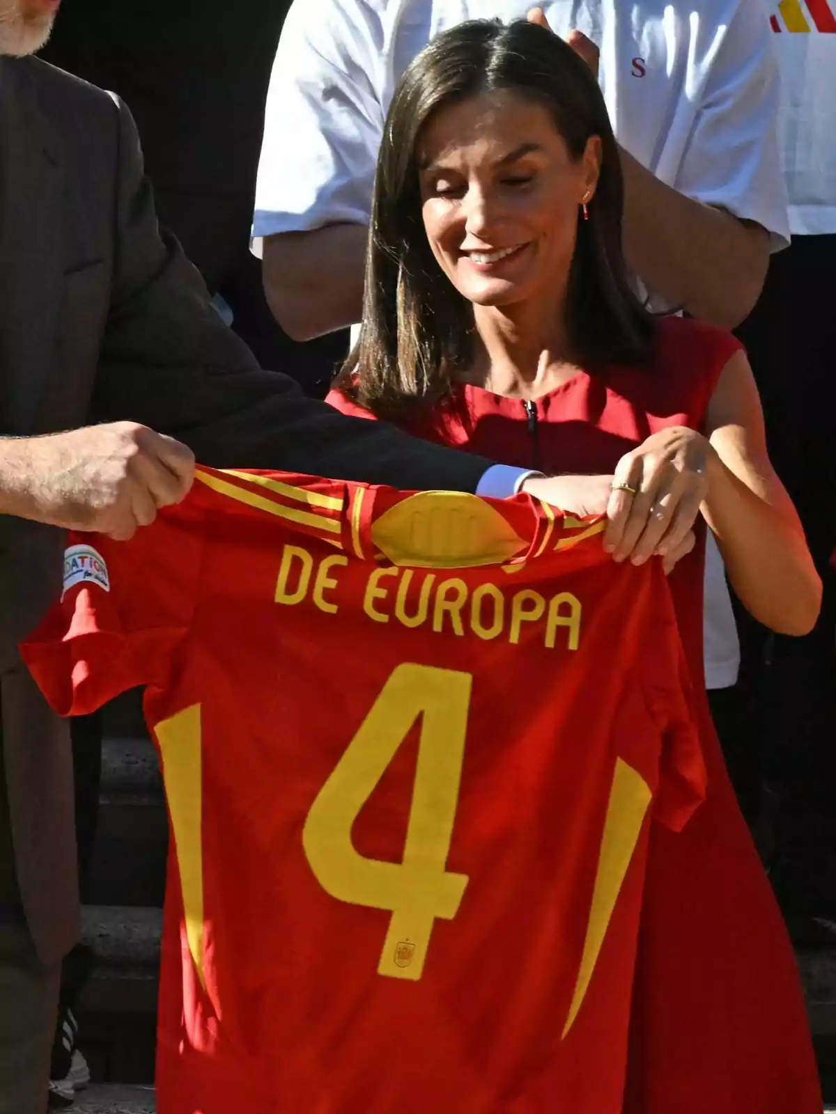 La reina, Letícia Ortiz, somrient, sosté una samarreta vermella de la selecció espanyola de futbol amb el número 4 i la frase La reina, Letícia Ortiz, somrient, sosté una samarreta vermella de la selecció espanyola de futbol amb el número 4 i la frase