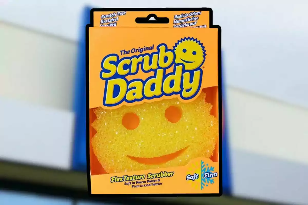 Esponja groga en forma de cara somrient dins d’un embolcall taronja amb el nom Scrub Daddy en lletres grans i blaves Esponja groga en forma de cara somrient dins d’un embolcall taronja amb el nom Scrub Daddy en lletres grans i blaves