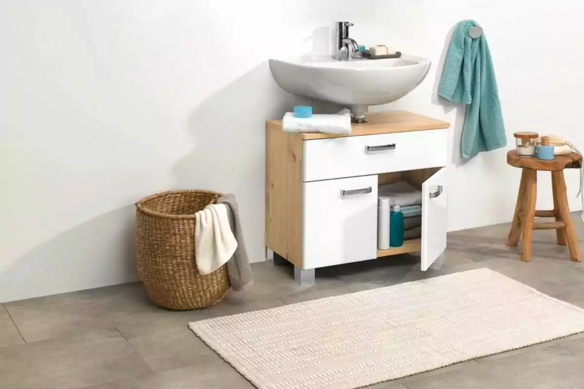 Bany modern amb lavabo blanc, moble de fusta, tovalloles, cistell de vímet i catifa clara sobre terra gris Bany modern amb lavabo blanc, moble de fusta, tovalloles, cistell de vímet i catifa clara sobre terra gris