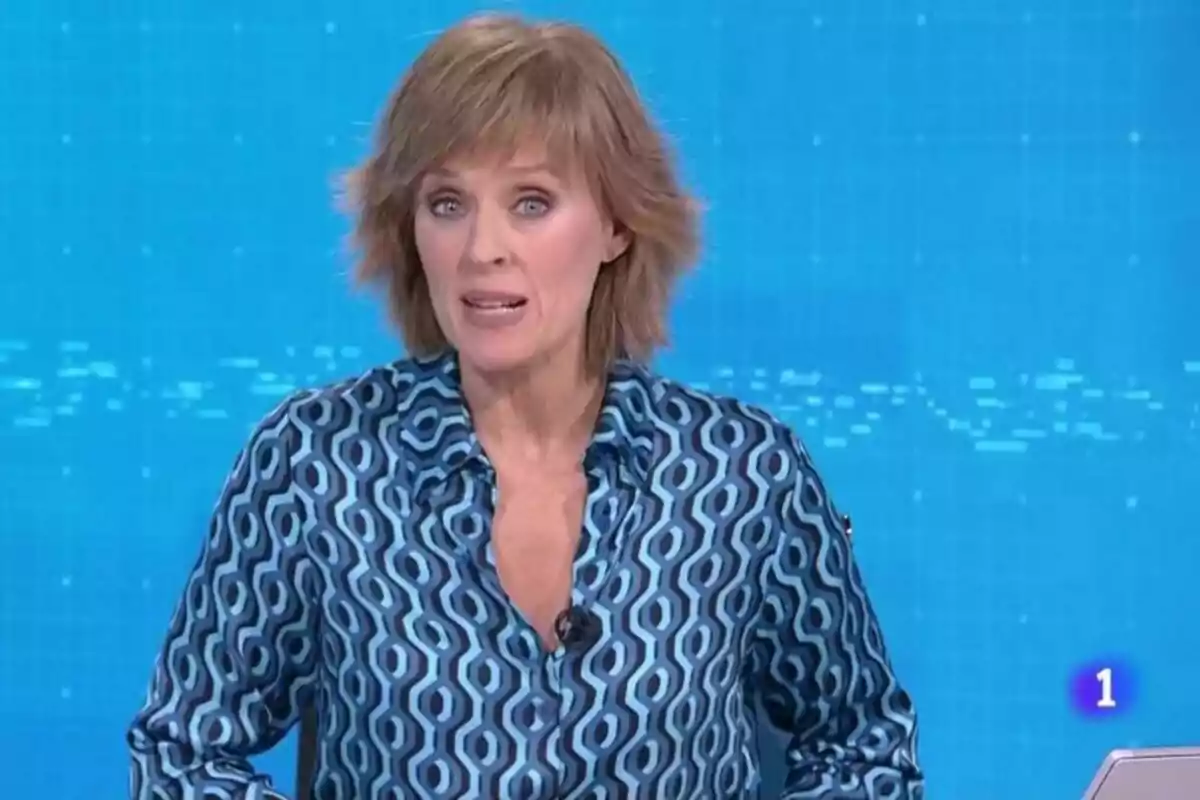 Marta Carazo, presentadora de noticias en RTVE, vestida con camisa azul de patrones geométricos frente a un fondo azul en un estudio de televisión. Marta Carazo, presentadora de noticias en RTVE, vestida con camisa azul de patrones geométricos frente a un fondo azul en un estudio de televisión.