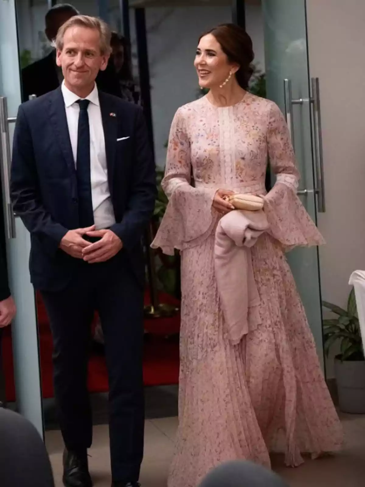 Un home amb vestit fosc i corbata al costat d’una dona amb un vestit llarg rosa clar i detalls florals, tots dos somriuen mentre caminen en un ambient elegant. Un home amb vestit fosc i corbata al costat d’una dona amb un vestit llarg rosa clar i detalls florals, tots dos somriuen mentre caminen en un ambient elegant.