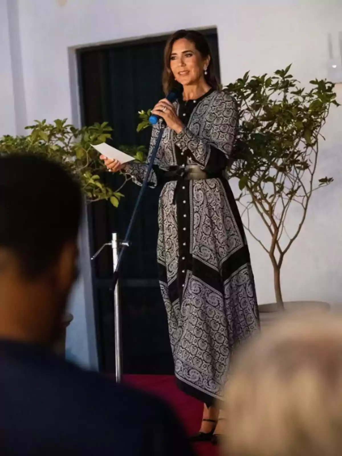 Dona dreta sobre una tarima parlant per micròfon i sostenint un full mentre porta un vestit llarg estampat en blanc i negre amb cinturó negre Dona dreta sobre una tarima parlant per micròfon i sostenint un full mentre porta un vestit llarg estampat en blanc i negre amb cinturó negre