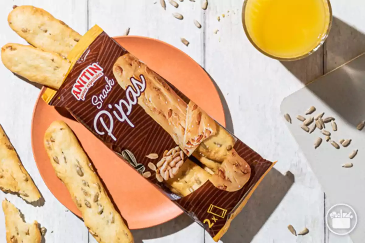 Paquet de snack de pipes Anitin sobre un plat taronja acompanyat de bastonets de pa, llavors de gira-sol i un got de suc de taronja Paquet de snack de pipes Anitin sobre un plat taronja acompanyat de bastonets de pa, llavors de gira-sol i un got de suc de taronja