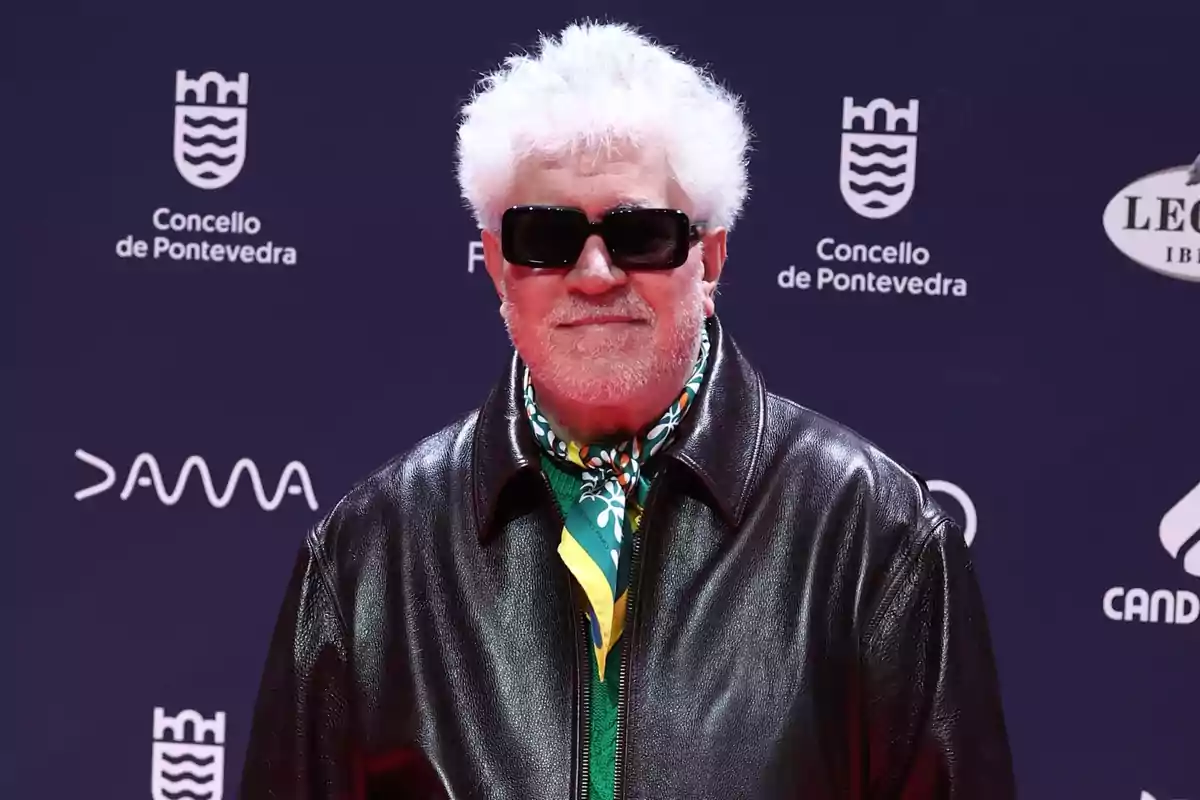 Pedro Almodóvar wearing dark sunglasses, a black leather jacket, and a colorful scarf, posing in front of a blue background with white logos. Pedro Almodóvar wearing dark sunglasses, a black leather jacket, and a colorful scarf, posing in front of a blue background with white logos.