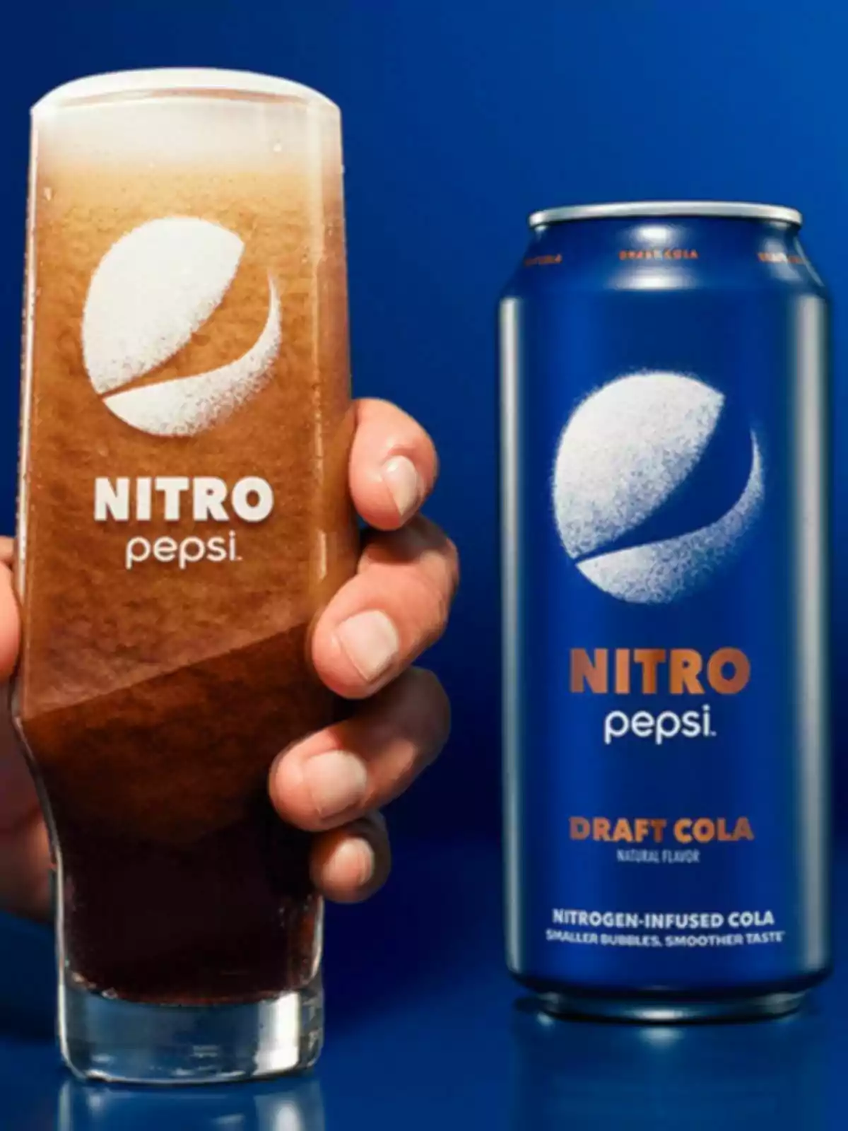 Una mano sostiene un vaso de Nitro Pepsi servido junto a una lata azul de la misma bebida sobre un fondo azul