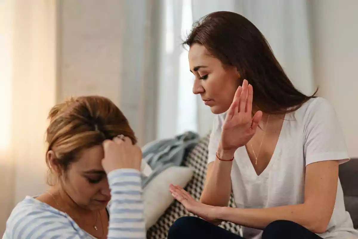 Dues dones assegudes en una habitació, una d’elles sembla molesta i aixeca la mà en senyal de rebuig mentre l’altra es mostra trista i capcotada.