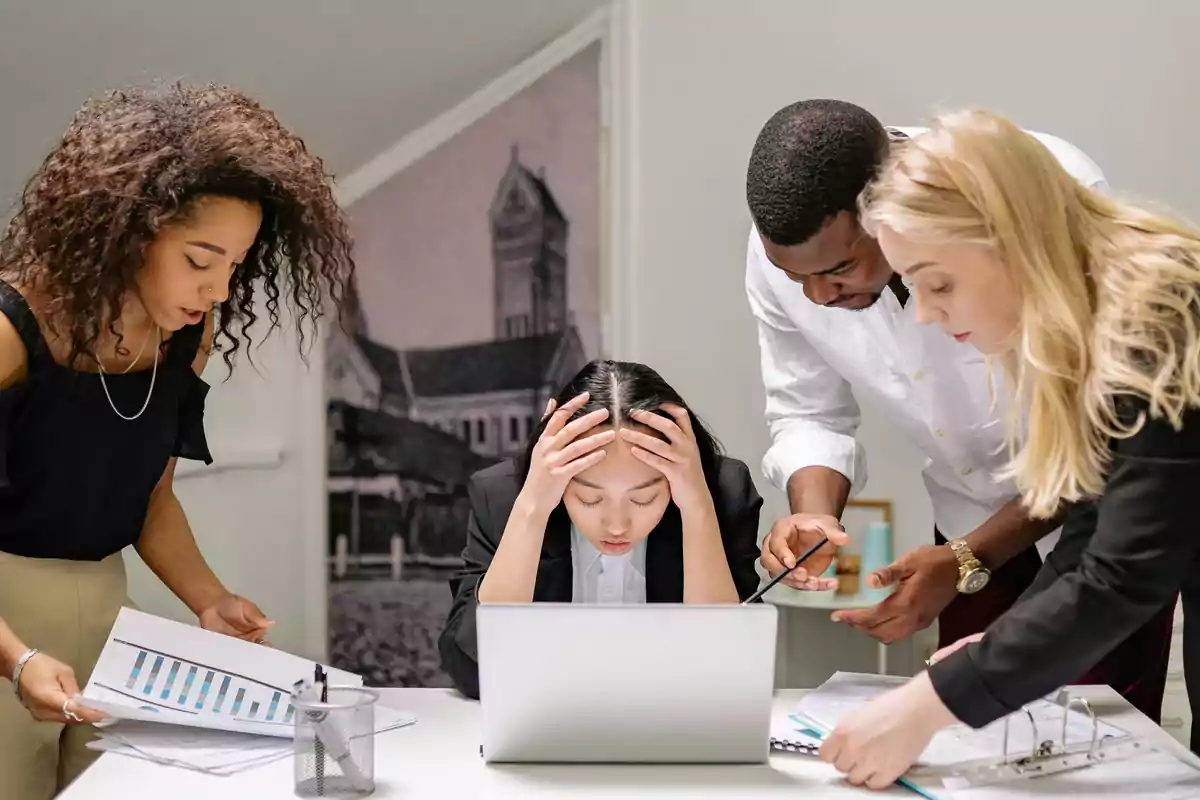 A woman sitting in front of a laptop holds her head in both hands, visibly overwhelmed, while her colleagues review documents. A woman sitting in front of a laptop holds her head in both hands, visibly overwhelmed, while her colleagues review documents.