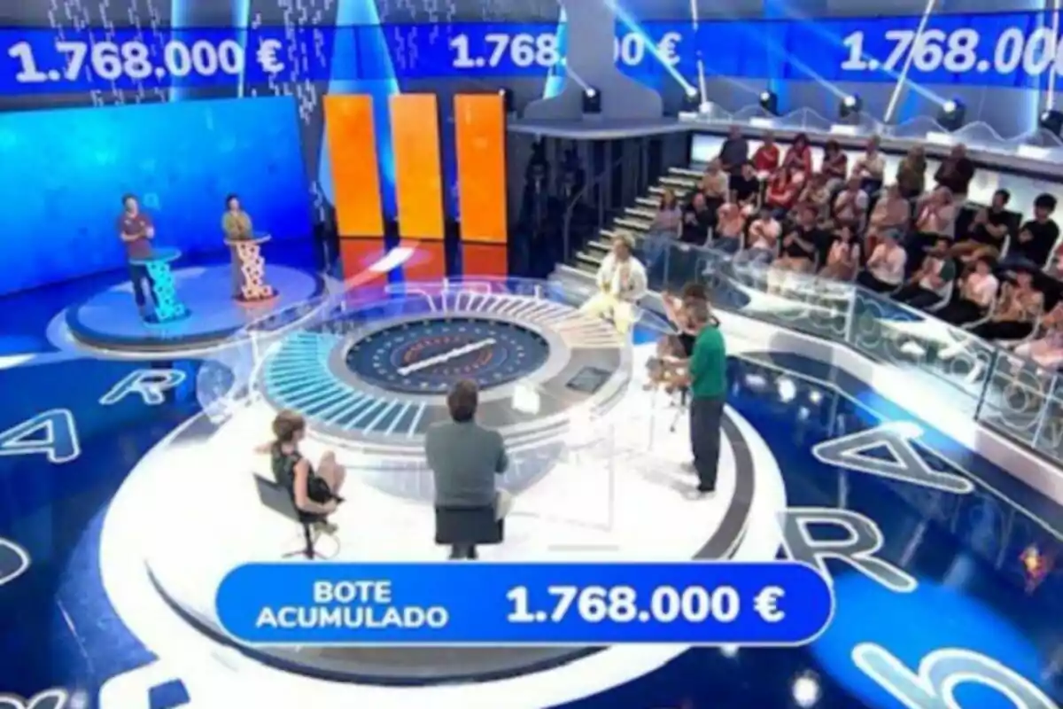 Pla general del plató de 'Pasapalabra' amb participants en un plató modern, públic observant i un premi acumulat d’un milió set-cents seixanta-vuit mil euros mostrat a la pantalla. Pla general del plató de 'Pasapalabra' amb participants en un plató modern, públic observant i un premi acumulat d’un milió set-cents seixanta-vuit mil euros mostrat a la pantalla.