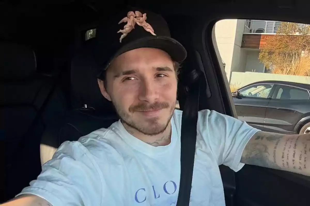 Brooklyn Beckham amb gorra negra amb disseny d’àngels assegut al seient del conductor d’un cotxe, amb samarreta blanca i somrient lleument. Brooklyn Beckham amb gorra negra amb disseny d’àngels assegut al seient del conductor d’un cotxe, amb samarreta blanca i somrient lleument.