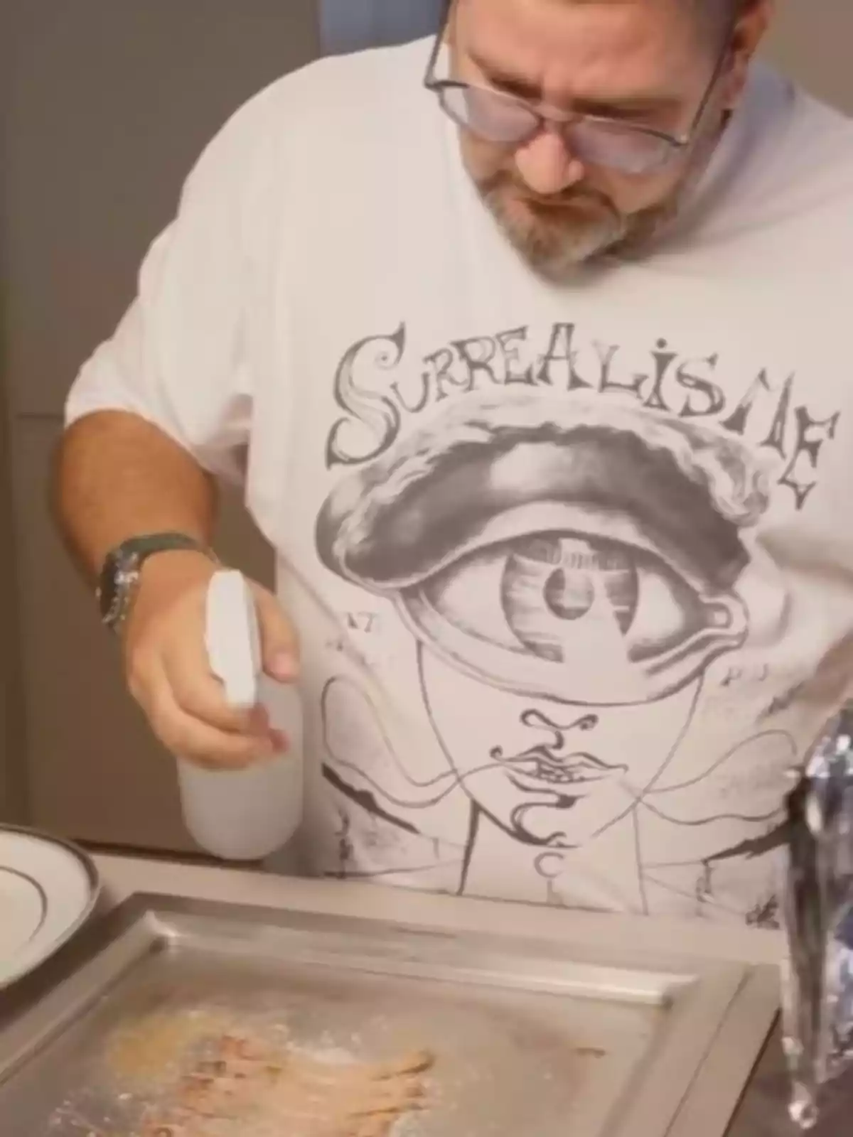 Man with glasses and a beard wearing a white t-jersey with a surreal design while cooking and holding a spray bottle over a griddle Man with glasses and a beard wearing a white t-jersey with a surreal design while cooking and holding a spray bottle over a griddle
