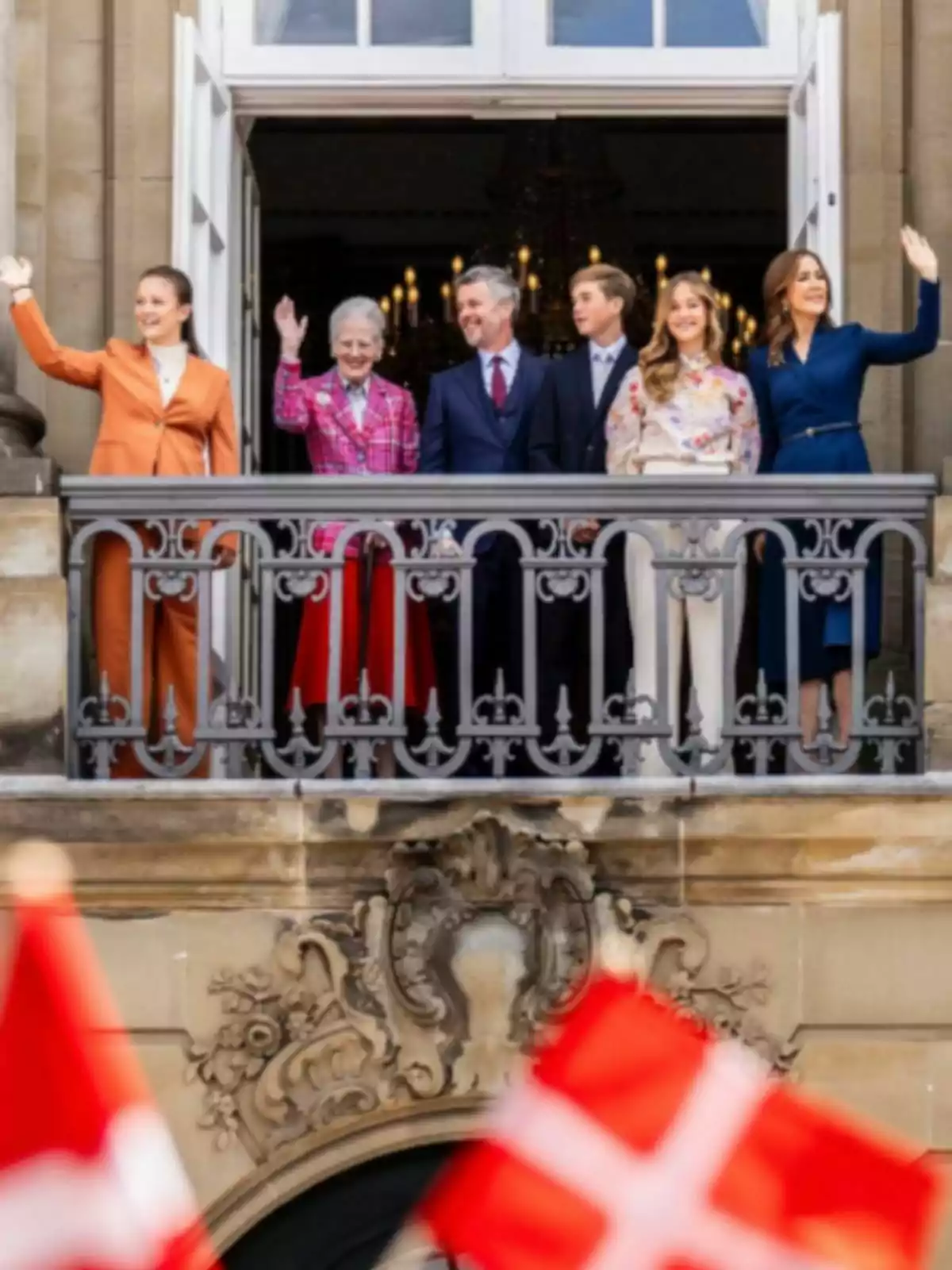 Seis personas elegantemente vestidas saludan desde un balcón decorado mientras varias banderas de Dinamarca ondean en primer plano Seis personas elegantemente vestidas saludan desde un balcón decorado mientras varias banderas de Dinamarca ondean en primer plano