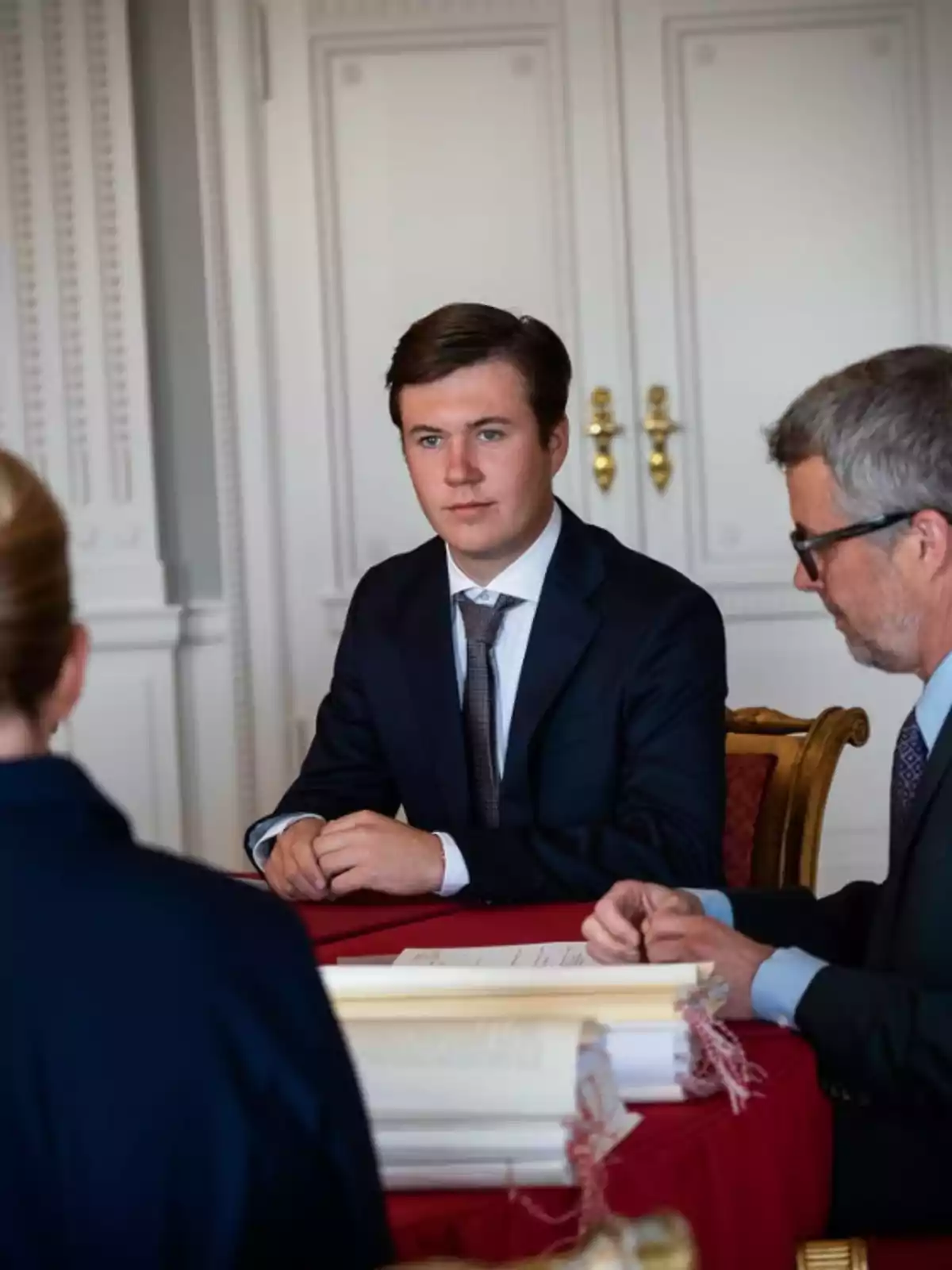 Joven con traje oscuro sentado en una mesa roja junto a dos personas en una sala elegante Joven con traje oscuro sentado en una mesa roja junto a dos personas en una sala elegante