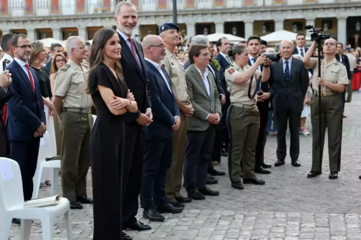 A group of people dressed in formal and military attire are standing in an outdoor plaza while watching and photographing an event. A group of people dressed in formal and military attire are standing in an outdoor plaza while watching and photographing an event.