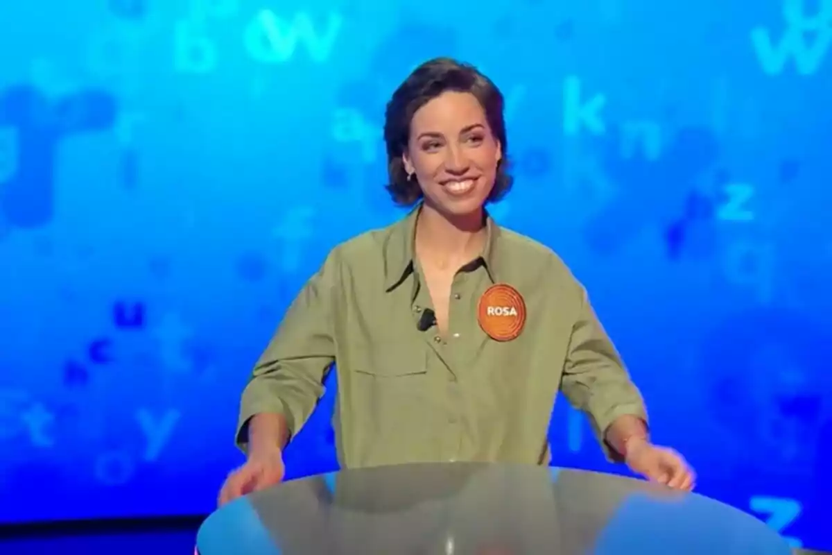 Rosa Rodríguez somrient amb una camisa verda i un identificador taronja que diu Rosa, participant en un programa de televisió, que és 'Pasapalabra', amb un fons blau ple de lletres. Rosa Rodríguez somrient amb una camisa verda i un identificador taronja que diu Rosa, participant en un programa de televisió, que és 'Pasapalabra', amb un fons blau ple de lletres.