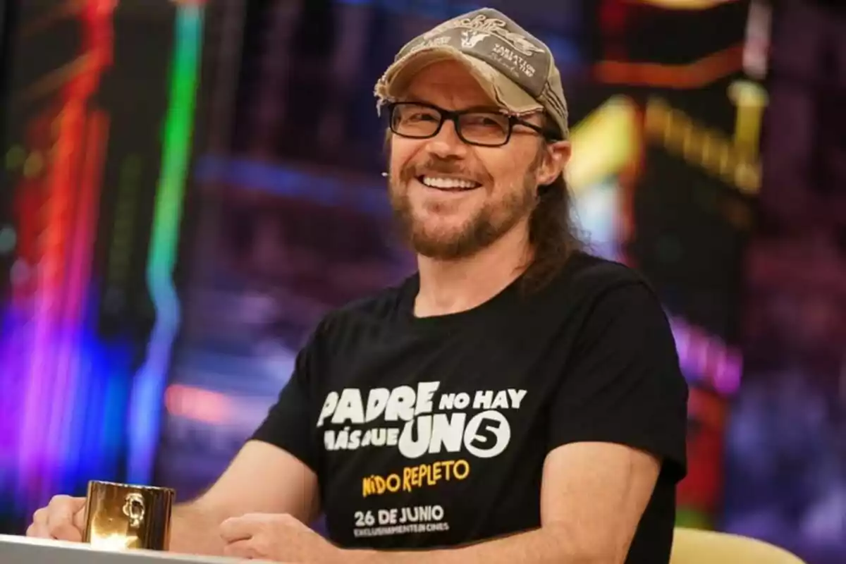 Santiago Segura, with a beard and glasses, smiles while sitting at a table. He is wearing a cap and a black t-jersey with white and yellow letters. The background is colorful and blurred on 'El Hormiguero'. Santiago Segura, with a beard and glasses, smiles while sitting at a table. He is wearing a cap and a black t-jersey with white and yellow letters. The background is colorful and blurred on 'El Hormiguero'.
