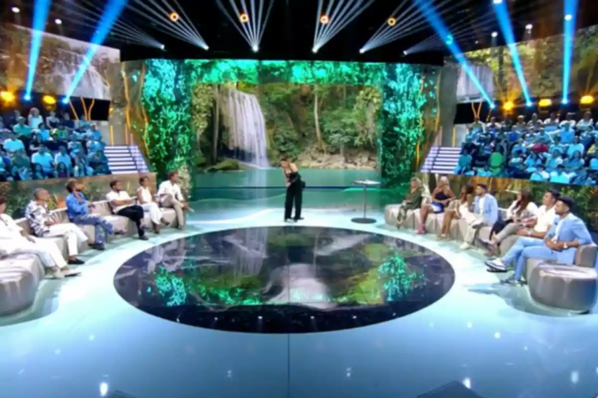 Un plató de televisió amb públic i participants asseguts en sofàs, una presentadora dreta al centre i una gran pantalla al fons mostrant una cascada envoltada de vegetació. Captura de 'Supervivientes' Un plató de televisió amb públic i participants asseguts en sofàs, una presentadora dreta al centre i una gran pantalla al fons mostrant una cascada envoltada de vegetació. Captura de 'Supervivientes'