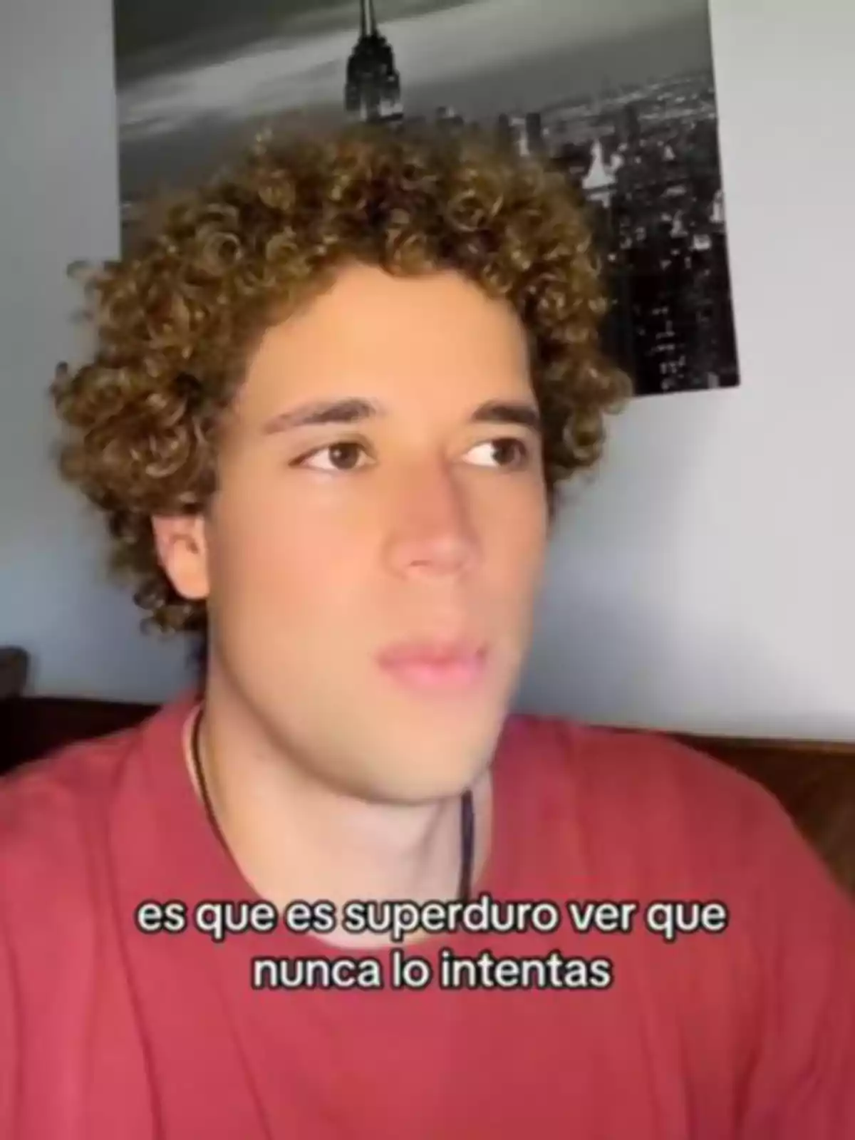 Young man with curly hair and a red t-jersey looking to the side with a black and white picture of a city in the background and text on the screen that says Young man with curly hair and a red t-jersey looking to the side with a black and white picture of a city in the background and text on the screen that says