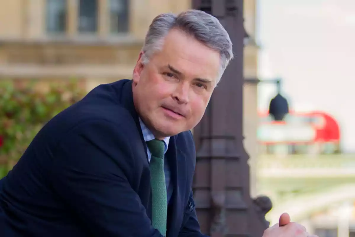 Tim Loughton in a blue suit and green tie posing outdoors in front of an old building Tim Loughton in a blue suit and green tie posing outdoors in front of an old building