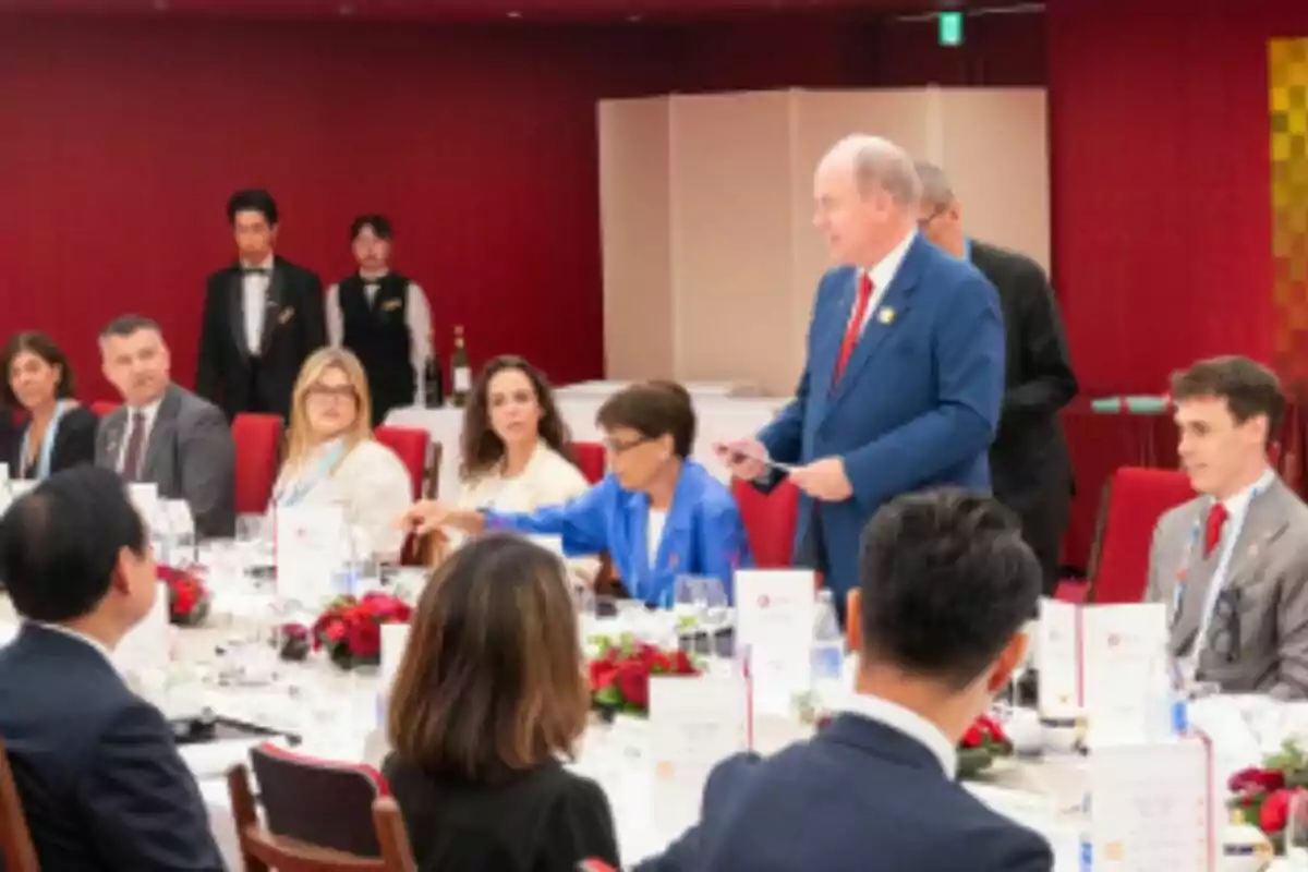 Un grup de persones elegantment vestides participa en un sopar formal en un saló decorat en tons vermells mentre un home amb vestit blau s’aixeca, que és l’Albert de Mònaco, i sembla que està fent un discurs al costat de l’Estefania de Mònaco. Un grup de persones elegantment vestides participa en un sopar formal en un saló decorat en tons vermells mentre un home amb vestit blau s’aixeca, que és l’Albert de Mònaco, i sembla que està fent un discurs al costat de l’Estefania de Mònaco.