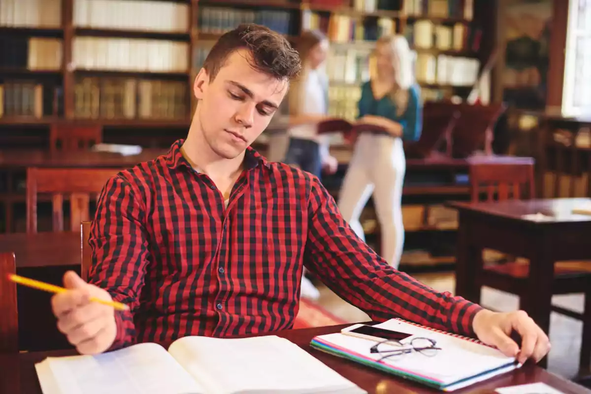 Joven con camisa de cuadros rojos estudiando en una biblioteca con libros abiertos y dos personas conversando al fondo