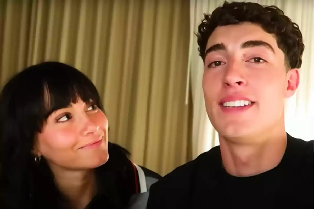 Aitana mira sonriente a Plex que está sonriendo frente a la cámara, con cortinas beige de fondo. Aitana mira sonriente a Plex que está sonriendo frente a la cámara, con cortinas beige de fondo.