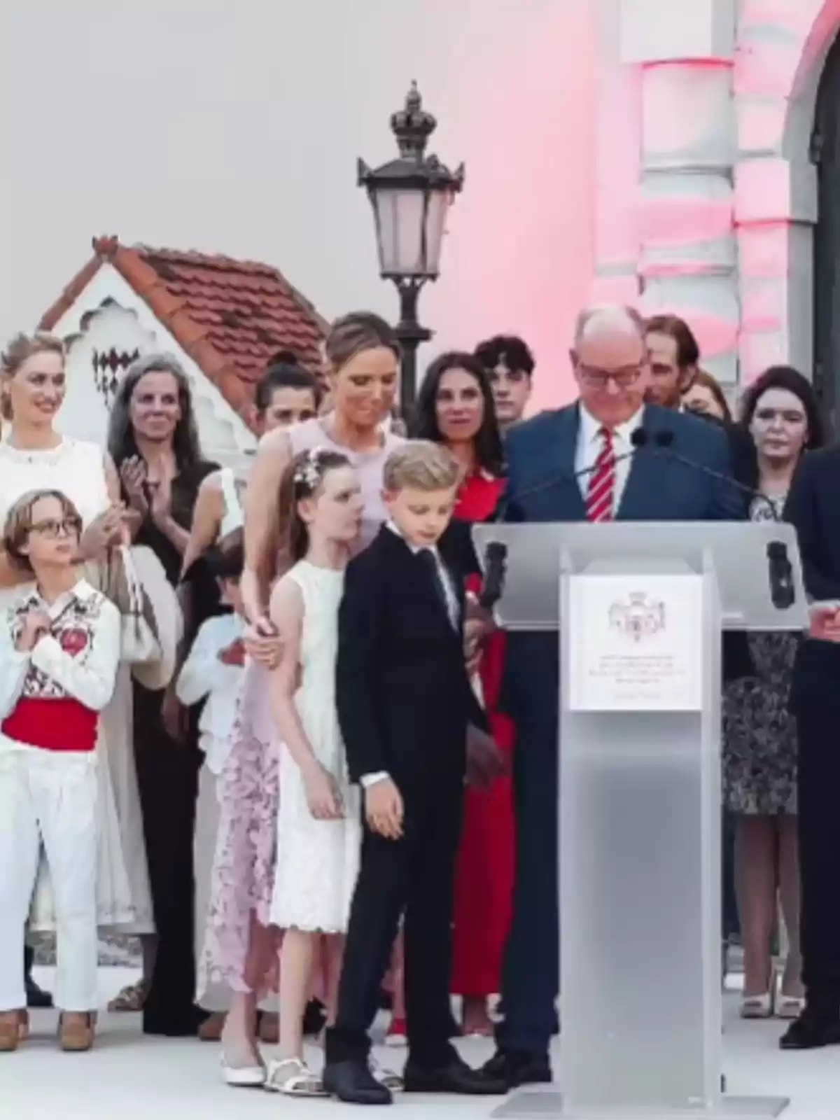 Un grup de persones elegantment vestides es reuneix al voltant d’un faristol en un esdeveniment a l’aire lliure, amb un home gran amb vestit i corbata parlant mentre nens i adults l’acompanyen. Un grup de persones elegantment vestides es reuneix al voltant d’un faristol en un esdeveniment a l’aire lliure, amb un home gran amb vestit i corbata parlant mentre nens i adults l’acompanyen.