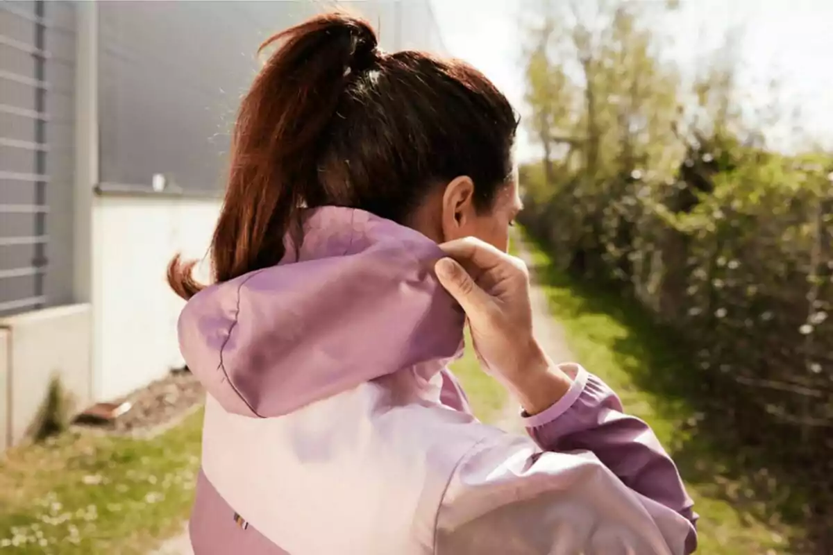 Mujer de espaldas con coleta ajustando la capucha de su chaqueta lila mientras camina al aire libre en un día soleado Mujer de espaldas con coleta ajustando la capucha de su chaqueta lila mientras camina al aire libre en un día soleado