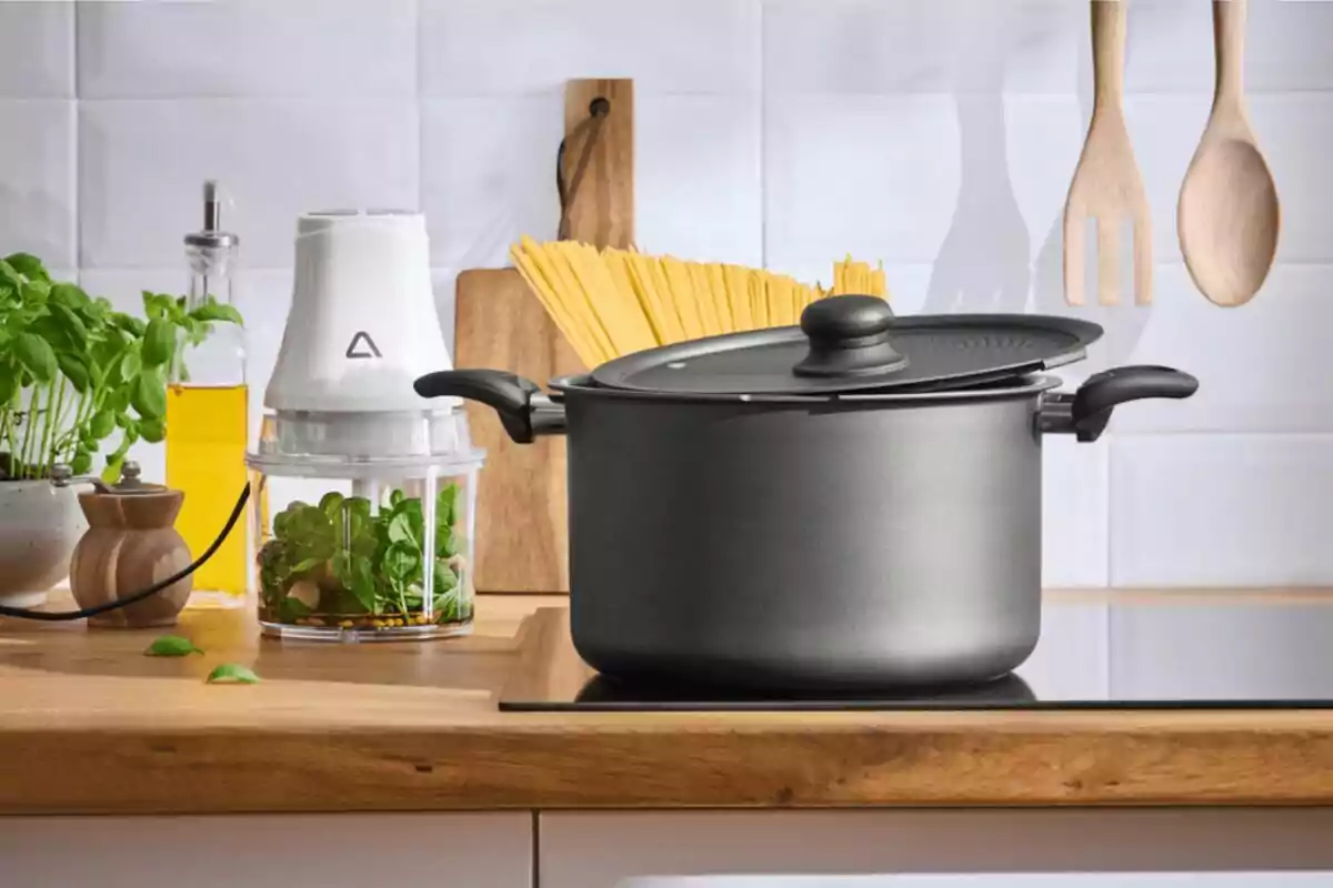 Olla negra amb tapa sobre una cuina en una cuina moderna amb utensilis de fusta, espaguetis crus, oli d’oliva, plantes i un processador d’aliments al fons.