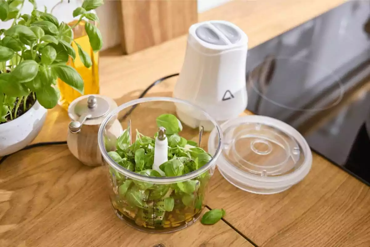 Processador d’aliments amb fulles fresques d’alfàbrega i altres ingredients en una cuina moderna sobre una taula de fusta
