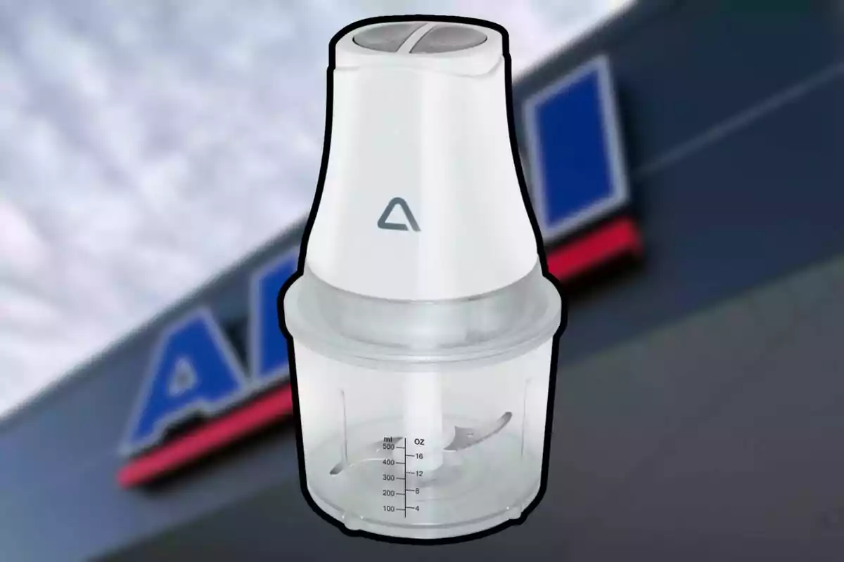 Processador d’aliments elèctric de color blanc amb recipient transparent i ganivetes d’acer inoxidable; al fons s’observa un rètol borros d’una botiga.