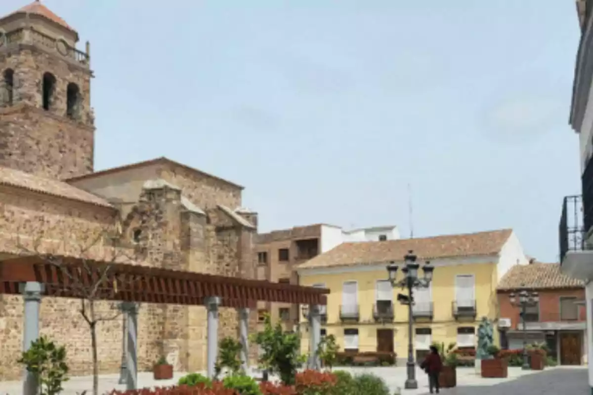 Plaça amb una església de pedra, cases de colors clars, una pèrgola de fusta i una persona caminant a Castella-la Manxa. Plaça amb una església de pedra, cases de colors clars, una pèrgola de fusta i una persona caminant a Castella-la Manxa.