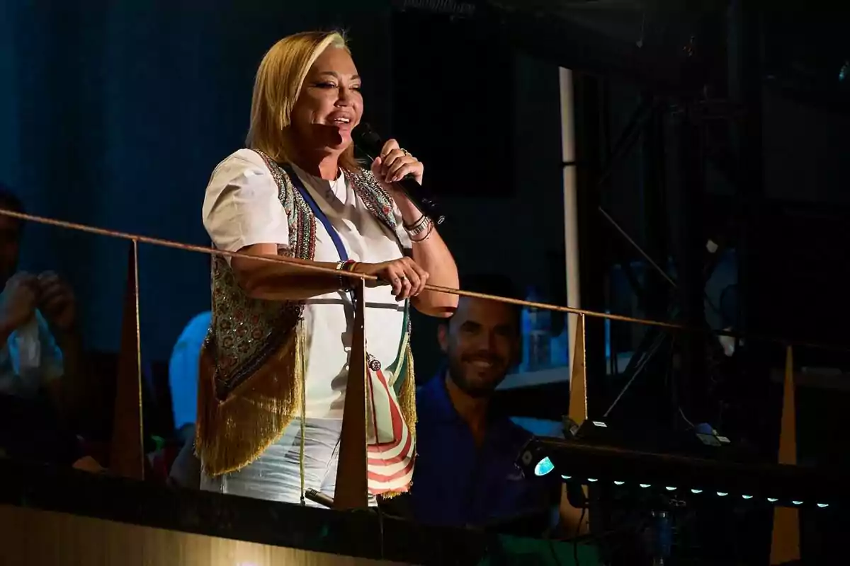 Belén Esteban somrient amb un micròfon a la mà, vestida amb una samarreta blanca i un xalec de colors, dreta en un balcó durant un esdeveniment nocturn. Belén Esteban somrient amb un micròfon a la mà, vestida amb una samarreta blanca i un xalec de colors, dreta en un balcó durant un esdeveniment nocturn.